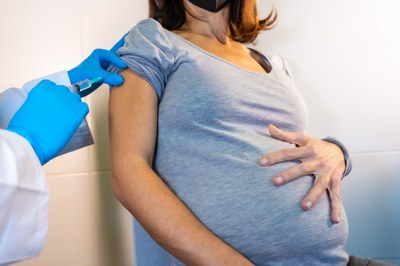 #ParaTodosVerem. Fotografia de mulher grávida usando camiseta cinza recebendo vacina de médico usando bata branca e luvas azuis.
