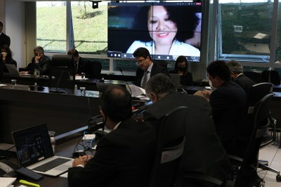 A foto é de uma mesa composta por conselheiros e, ao fundo, uma TV exibe a imagem de Cláudia Pinho, que participa por videoconferência