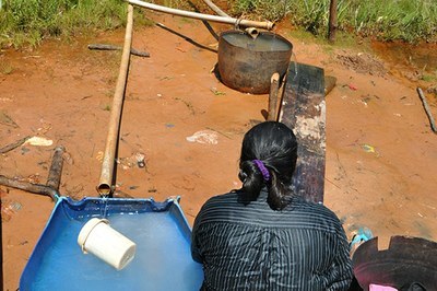 #Pracegover Fotografia em que uma pessoa indígena é vista de costas. Ao lado e à frente dela, dois galões com encanamento improvisado armazenam água.