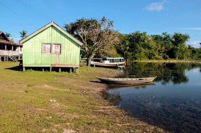Casa de madeira ao lado de um lago e de uma árvore. Há dois barcos na beira do lago. Floresta ao fundo. Dia ensolarado.