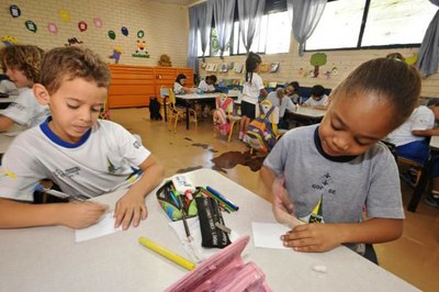 Duas crianças em mesa escolar fazem tarefa em sala de aula. Ao fundo, outros estudantes.