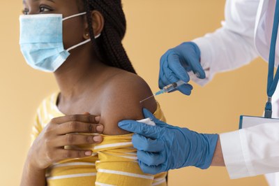 #pracegover - a imagem mostra uma mulher jovem negra sendo vacinada contra a covid19. Ela usa uma máscara de proteção no rosto.