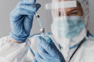 #Pracegover Foto de um profissional da saúde, paramentado com EPIs manipulando uma seringa e um frasco de vacina 