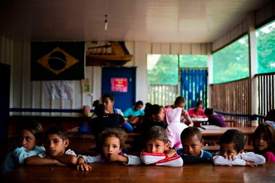 A imagem mostra crianças sentadas em uma sala de aula de uma escola no campo.