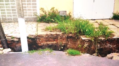 Foto mostra uma cratera formada em um chão de asfalto, próximo a calçadas que passam na frente de casas. O buraco é grande e com terra aparente.