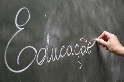Arte retangular sobre foto de quadro escolar com uma pessoa escrevendo a palavra educação com giz branco.