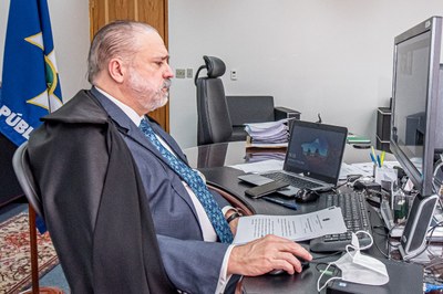 Foto do pgr sentado em frente ao computador, participando da sessão do STF. ele está usando terno azul e capa preta por cima.