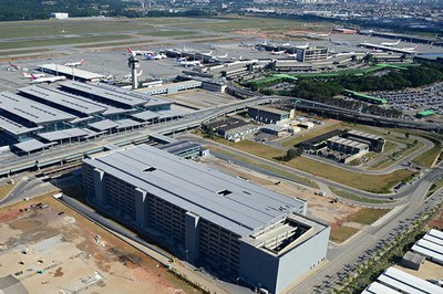 Na imagem, se tem a vista aérea do Aeroporto Internacional de Guarulhos, com os terminais, as construções e, ao fundo, a pista de aviões.