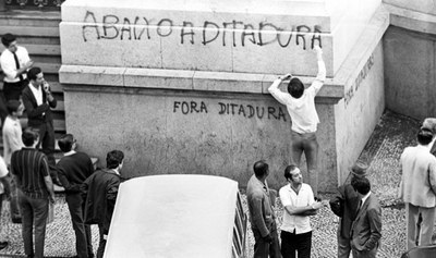 #pracegover: Foto preta e branca mostra pessoas em torno de uma parede em que se lê "Abaixo a Ditadura" e "Fora Ditadura". A foto é de Kaoru do CPDoc.