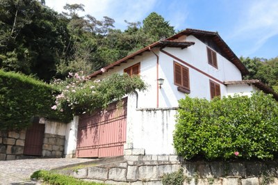 #pracegover: fachada do imóvel que ficou conhecido como "Casa da Morte", uma casa branca, de janelas marrons, cercada por árvores e plantas
