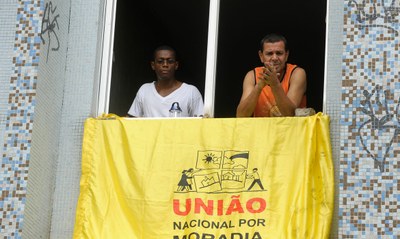#pracegover: dois homens na janela de um prédio, onde está pendurada uma faixa amarela na qual se lê "União Nacional por Moradia"