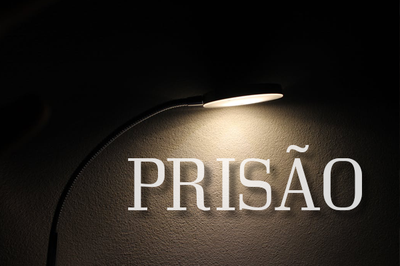 #pracegover: imagem retratando uma luminária sobre um fundo escuro e, na luz da luminária, a palavra prisão em fonte branca 