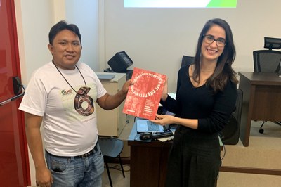 Foto mostra procuradora da República Manoela Lopes Lamenha Lins Cavalcante recebendo de líder indígena Dário Kopenawa documento de capa vermelha.