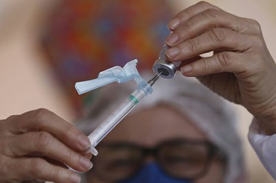 Mulher com touca e máscara cirúrgica segura seringa e vidro de vacina em frente aos olhos. 