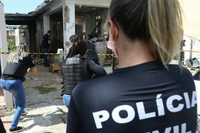 Mulher loira, de costas, com camiseta preta com a palavra "polícia" observa duas outras mulheres policiais entrarem em um imóvel.