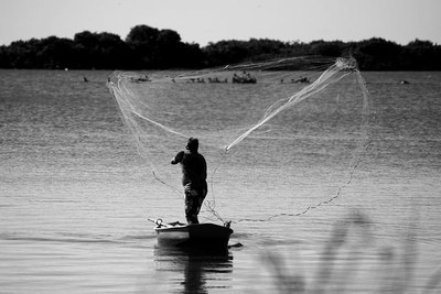 Registro fotográfico em preto de branco de pescador em pé numa canoa jogando sua rede na água