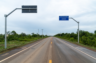 #PraTodosVerem: Imagem da rodovia BR-319. É a divisa dos estados do Amazonas e Rondônia com uma rua pavimentava cercada por mato e duas placas.