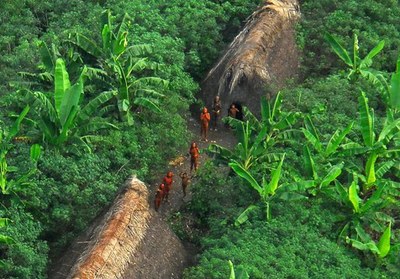 Foto aérea de trecho de floresta com duas ocas e alguns índios isolados, ao longe