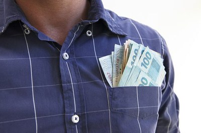 Homem de camisa azul, sem mostrar o rosto, com várias notas de reais no bolso