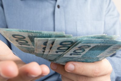 #PraCegoVer Fotografia de uma pessoa segurando algumas notas de cem reais. O foco da imagem está no dinheiro e a pessoa ficando ao fundo. 