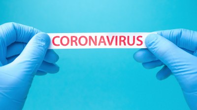 Imagem de duas mão usando luvas azuis segurando papel branco onde se lê em letras vermelhas a palavra Coronavirus