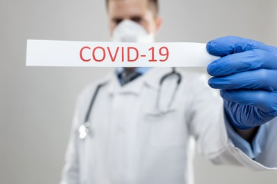 Foto tirada em fundo branco mostra um médico segurando em destaque pedaço de papel onde se lê na cor vermelha a palavra Covid-19