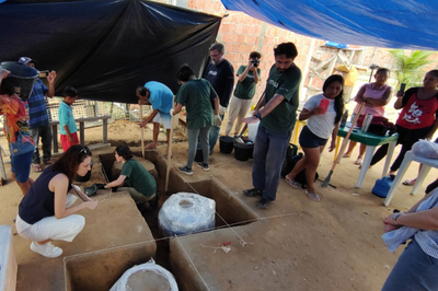 A foto mostra três urnas em formato cilíndrico embaladas sendo retiradas do sítio arqueológico. Há várias pessoas em volta observando os trabalhos 
