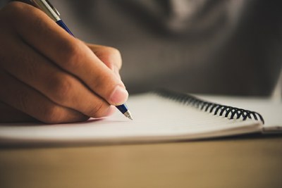 Mão segurando uma caneta em posição de escrever em um caderno.
