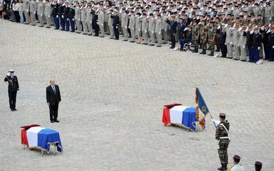 foto mostra cerimônia de homenagem aos policiais mortos em emboscada.Dois caixões, cobertos com a bandeira da França, estão no meio de um pátio. Ao redor, vários militares fardados estão de pé.