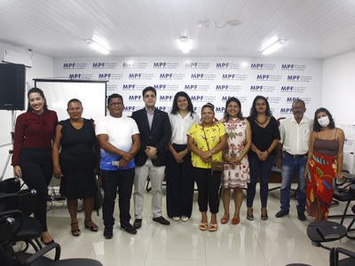 foto mostra participantes da reunião, de pé, posando para foto. AO fundo, painel com logomarca do MPF.
