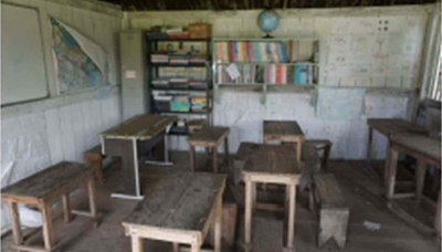 Foto lateral de sala de aula com mesas e cadeiras em madeira, quadro branco, chão de terra batida, com duas estantes de livros ao fundo. Há um globo terrestre sobre uma das estantes