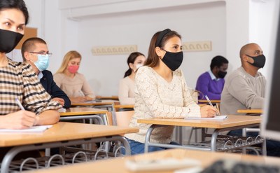 Foto mostra alunos adultos em sala de aula, usando máscaras e sentados com distância entre as carteiras