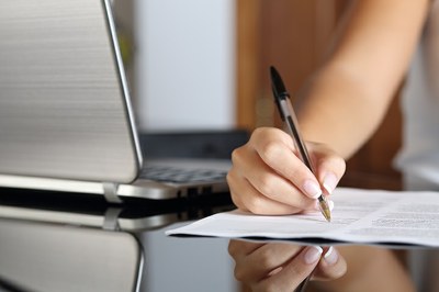 #pracegover foto mostra mulher fazendo anotações com caneta preta em uma folha de papel. ao lado há um notebook
