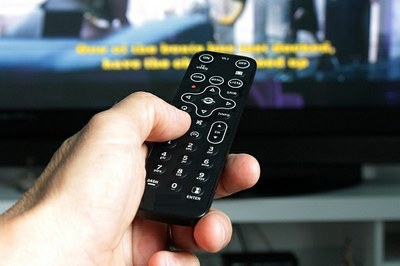 Foto retangular de Mão masculina apontando o controle remoto para a TV onde há legendas