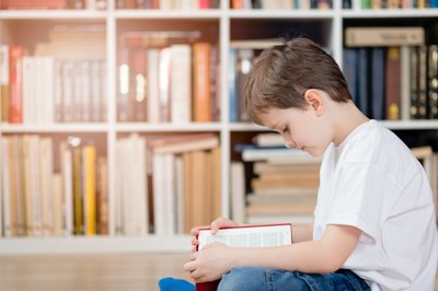Criança sentada no chão lendo um livro, com um estante cheia de outros livros ao fundo.