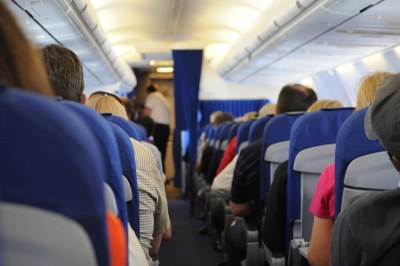 Interior de um avião com passageiros sentados nas poltronas, de costas para o observador, e um comissário de bordo ao fundo do corredor