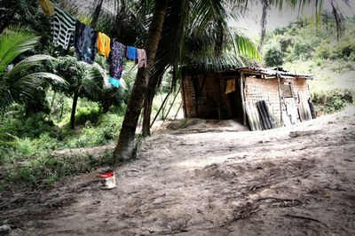 Foto da comunidade quilombola Rio dos Macacos mostrando uma pequena casa de estrutura rústica e frágil e um varal com algumas roupas penduradas.