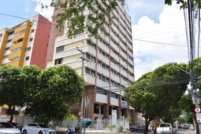 Foto do edifício da Procuradoria da República no Ceará. Ao redor do prédio há arvores. E na rua carros estacionados e em circulação.