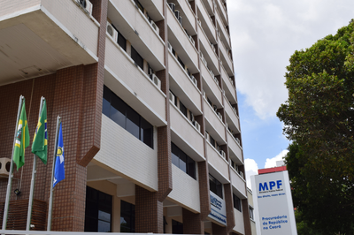 Foto lateral do prédio da sede do MPF em Fortaleza. Em primeiro plano aparece uma placa com nome da instituição e bandeiras do Brasil, do Ceará e do Ministério Público. 