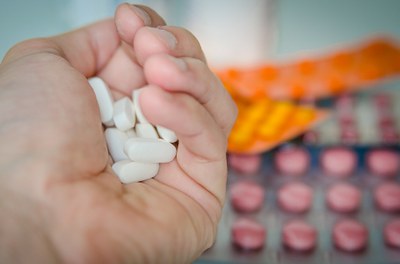 Foto que mostra em primeiro plano uma mão cheia de comprimidos e em segundo plano diversas cartelas de comprimidos.