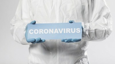 Pessoa com roupa branca de proteção com uma placa na mão onde se lê escrito coronavírus