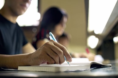 Foto mostra a mão de um estudante escrevendo em um caderno com uma lapiseira em primeiro plano e outros estudantes ao fundo, com imagem desfocada