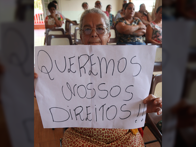 Fotografia tirada no dia da audiência mostra uma senhora idosa sentada no auditório segurando um cartaz na qual está escrito "Queremos nossos direitos".