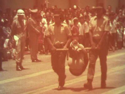 Cena tirada de um documentário mostra um índio carregado em um instrumento de tortura conhecido pelo nome de pau de arara, com as mãos e pés amarrados, sendo carregado por soldados em um desfile.
