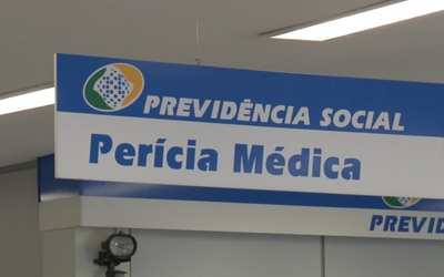 Imagem mostra parte de um letreiro interno de agência da Previdência onde se lê Previdência Social - Perícias. A placa tem o fundo branco e as letras são azuis.
