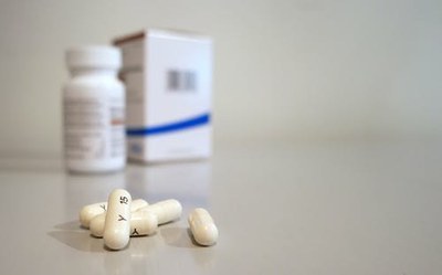 Fotografia ilustrativa que mostra em primeiro plano algumas pílulas de remédio, e ao fundo, desfocado, a caixa de um remédio sem identificação.
