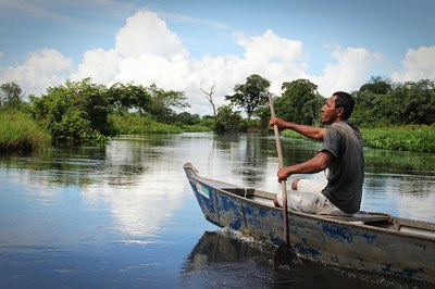 A foto mostra um homem conduzindo uma pequena canoa de madeira com um remo em um rio com vasta vegetação nas duas margens.