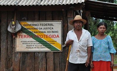 Na foto vê-se um casal de indígenas, vestidos com roupa simples de agricultores, em frente a um barraco e uma placa que diz Terra Protegida