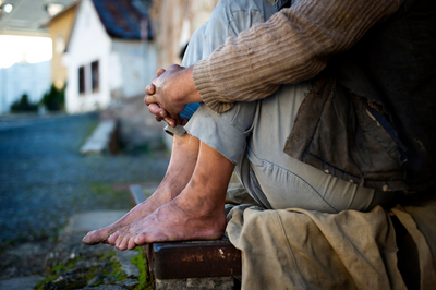 A foto mostra uma pessoa sentada na rua, abraçando as próprias pernas, com as roupas, os pés e as mãos bastante sujos. O enquadramento da foto exclui o rosto da pessoa.