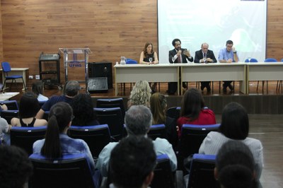 Foto que mostra a mesa principal do evento em uma das palestras e detalhe do público de costas.